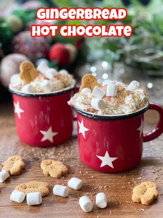Two mugs of gingerbread hot chocolate in red enamel mugs with stars, topped with whipped cream, grated chocolate, marshmallows and mini gingerbread men.