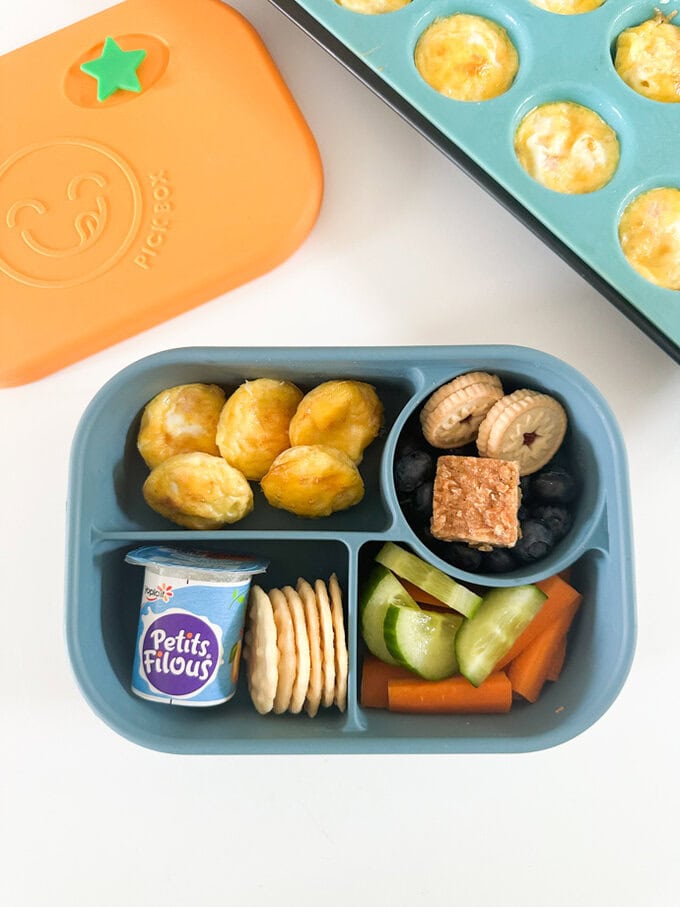 Five Mini egg bites arranged in a child's lunchbox, along with fresh chopped veggies,fruit and sweet treats.