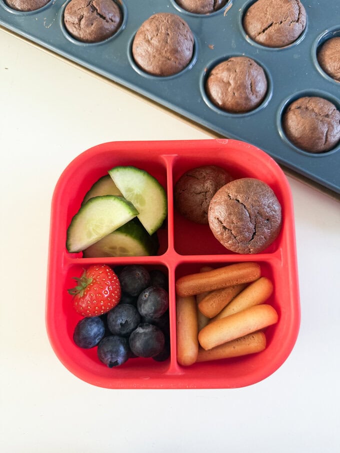 Two mini muffins served in a bright red divided,square snack box along with fresh fruit,cucumber slices and mini bread sticks.