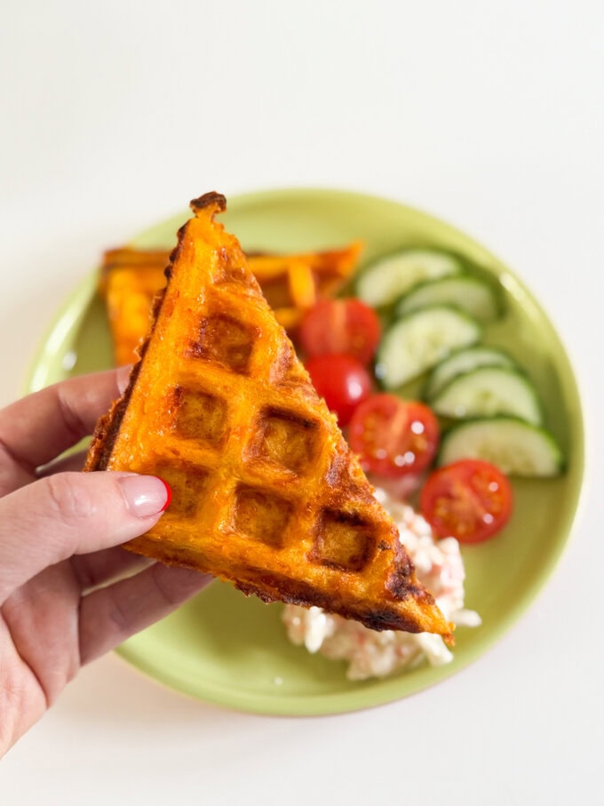 Cheesy Carrot Waffles served on a round plate along with sliced cherry tomatoes and cucumber and a dollop of coleslaw.