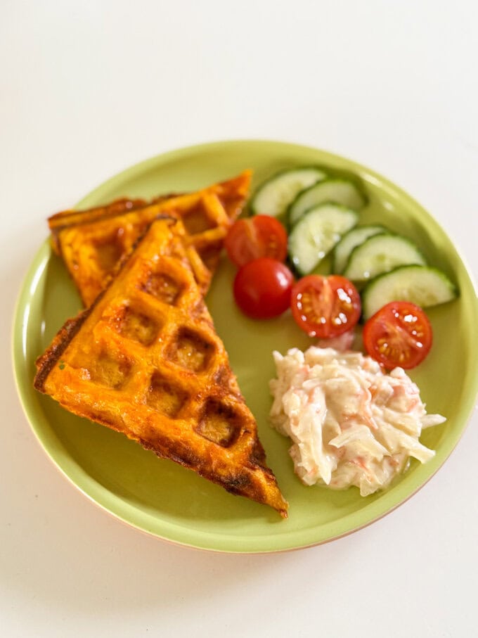 Cheesy Carrot Waffles served on a round plate along with sliced cherry tomatoes and cucumber and a dollop of coleslaw.